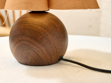 Load image into Gallery viewer, A close-up of a table lamp with a round dark brown wooden base and a beige fabric shade, sitting on a light textured surface with a black power cord visible.
