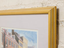 Load image into Gallery viewer, A close-up of a framed art print showing a colorful street scene with brick buildings and a blue sky, featuring a prominent gold frame and white matting against a textured white brick wall.
