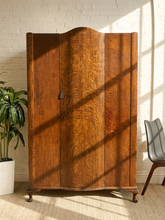Load image into Gallery viewer, Antique dark wood armoire with figured grain, tall curved center door, arched top, decorative metal pull, and rounded feet

