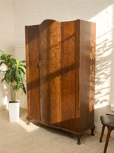 Load image into Gallery viewer, A tall vintage wooden wardrobe with three doors and a curved top, made of rich brown wood with visible grain, set against a white brick wall with sunlight and leaf shadows.
