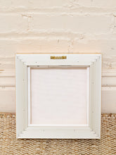 Load image into Gallery viewer, A square white wooden picture frame with a blank white center, photographed against a light beige painted brick wall and woven jute rug.
