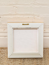 Load image into Gallery viewer, A square white wooden picture frame with a gold metal hanger at the top, shown empty against a white brick wall and a woven beige rug.
