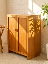 Load image into Gallery viewer, A mid-century modern wooden cabinet with two doors and vertical handles, styled in a room with a white brick wall, sunlight, and a potted plant.
