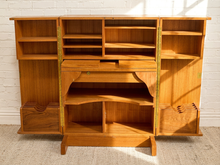 Load image into Gallery viewer, A mid-century modern wooden writing desk with integrated shelving, shown open against a white brick wall.
