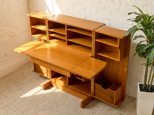 Load image into Gallery viewer, A mid-century modern wooden desk and shelving unit with a warm honey finish, set against a white brick wall with a potted plant on a beige carpet.
