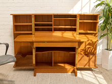 Load image into Gallery viewer, A mid-century modern wooden writing desk with a hutch and open shelving, shown in a bright room with sunlight casting leaf shadows on the wood and a potted plant in the background.

