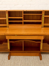 Load image into Gallery viewer, A warm medium-brown wooden writing desk with an open hutch and pull-out legs, set against a white brick wall and light beige carpet.
