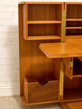 Load image into Gallery viewer, A close-up of a wooden writing desk cabinet with a drop-down writing surface, open shelves, a lower storage compartment, and brass hinges, set against a white brick wall.
