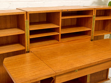 Load image into Gallery viewer, A mid-century modern wooden writing desk with an attached open shelving unit, made of warm orange-brown wood with brass-colored hinges.
