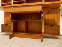 Load image into Gallery viewer, A close-up of a wooden writing desk with brass hardware, shown open to reveal shelves and drawers, set against a light brick wall and textured rug.

