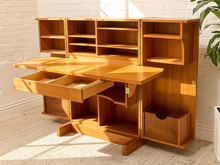 Load image into Gallery viewer, A mid-century modern wooden writing desk with a hutch, featuring a warm honey-amber finish, brass hardware, and an open drawer on the left, set against a white brick wall and beige carpet.
