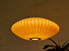 Load image into Gallery viewer, A warm, glowing paper lantern pendant light hanging against a textured brick wall, casting a soft golden light.
