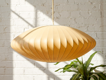 Load image into Gallery viewer, A large, organic-shaped pendant light made of light natural wood with a ribbed, pleated texture, hanging against a white brick wall with soft window shadows and a green plant in the foreground.
