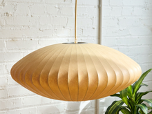 Load image into Gallery viewer, A mid-century modern pleated paper shade pendant light in a warm tan color, hanging against a white brick wall with a green plant visible in the lower right corner.
