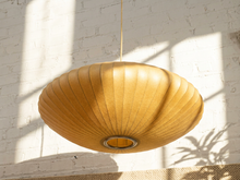 Load image into Gallery viewer, A large, round, saucer-shaped pendant light made of pleated natural wood, suspended from a thin cord against a white brick wall with dramatic window shadows.
