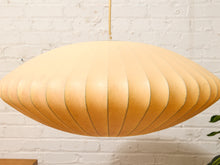 Load image into Gallery viewer, A large, mushroom-shaped pendant light with a pleated paper or fabric shade in a warm beige or light tan color, hanging against a white brick wall.
