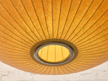 Load image into Gallery viewer, A close-up view of a warm yellow-orange paper lantern ceiling light fixture, showing its radial pleated structure and circular opening against a white brick wall background.
