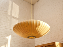 Load image into Gallery viewer, A large, disc-shaped pendant light with a pleated, fan-like structure in a warm natural wood finish, suspended from a thin cord in a room with white brick walls.
