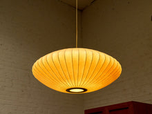 Load image into Gallery viewer, A warm, glowing paper lantern pendant light hanging from a thin cord in a room with a white brick wall.
