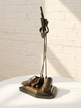 Load image into Gallery viewer, A bronze sculpture of golf clubs and shoes on a base, displayed on a white table against a white brick wall with sunlight and shadows.
