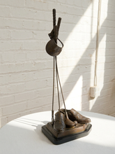 Load image into Gallery viewer, A decorative sculpture or display stand featuring a pair of brown leather shoes on a black base, with a hanging metal loop holding a small object, set on a white table against a white brick wall in bright natural light.
