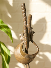 Load image into Gallery viewer, A rustic bronze-toned metal wall hook shaped like a stylized hand holding a spiral staff, shown against a warm off-white brick wall with leaf shadows and green leaves at the left edge.
