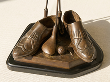 Load image into Gallery viewer, Bronze-colored metal golf-themed sculpture of a pair of shoes with golf clubs and a golf ball, displayed on a black base against a light beige textured background.
