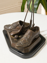 Load image into Gallery viewer, A decorative bronze sculpture of a pair of golf shoes on a black base, with golf clubs and a golf ball integrated into the design, displayed on a white tablecloth with a blurred indoor background.
