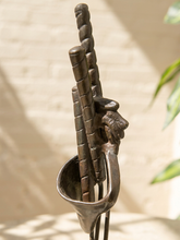 Load image into Gallery viewer, A close-up of a dark bronze metal fireplace tool set, including a poker, brush, and chisel, held in a matching cup holder against a warm beige brick wall background.
