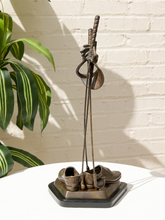 Load image into Gallery viewer, A decorative bronze sculpture of golf clubs and shoes on a black base, displayed on a white table against a white brick wall with a green plant leaf on the left.
