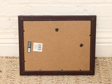Load image into Gallery viewer, The back of a dark brown wooden picture frame with a brown cardboard backing, showing mounting holes and a product label, set against a white brick wall and woven rug.
