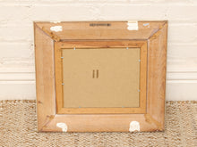 Load image into Gallery viewer, The back of a square wooden picture frame with a natural light brown finish, showing the backing board and corner protectors, resting on a woven rug against a white brick wall.
