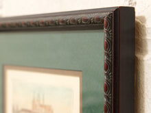 Load image into Gallery viewer, Close-up of a dark brown ornate picture frame with a decorative carved border and a sage green mat, holding a framed print against a textured white wall.
