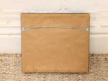 Load image into Gallery viewer, The back of a square framed picture with a brown backing board and a metal hanging wire with clips, resting on a woven rug against a white brick wall.
