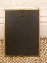 Load image into Gallery viewer, The back of a rectangular picture frame with a gold-toned border and a hanging loop, resting on a woven mat against a white paneled wall.
