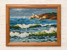 Load image into Gallery viewer, Vintage framed needlepoint coastal scene with sailboat, deep blue water, rocky shoreline, green landscape, and warm wood frame
