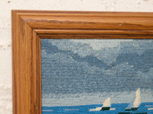 Load image into Gallery viewer, Close-up of a framed needlepoint canvas showing a blue ocean and sky with sailboats, bordered by a medium brown wooden frame against a white textured wall.
