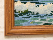 Load image into Gallery viewer, Close-up of a framed needlepoint canvas showing a landscape design with blue water, green foliage, and cream clouds, set in a warm brown wooden frame against a textured off-white wall.
