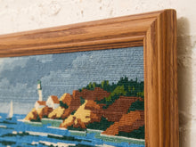 Load image into Gallery viewer, Close-up of a framed needlepoint canvas featuring a coastal landscape with blue water, brown and green hills, and a lighthouse, set in a warm wood frame against a textured off-white wall.
