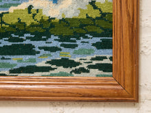 Load image into Gallery viewer, Close-up of a framed needlepoint canvas featuring a landscape design with green trees, blue and white water, and a warm brown wooden frame against a textured off-white wall.
