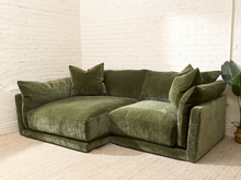 Load image into Gallery viewer, An olive green velvet sectional sofa with matching throw pillows, set in a room with white brick walls and a light beige carpet.
