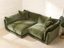 Load image into Gallery viewer, An olive green velvet sectional sofa with matching throw pillows, set in a bright room with white brick walls and a light beige carpet.
