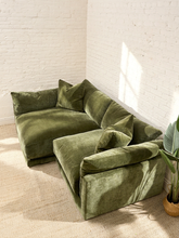 Load image into Gallery viewer, An olive green velvet sectional sofa with matching throw pillows, styled in a bright room with white brick walls, a beige rug, and a potted plant.
