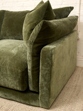 Load image into Gallery viewer, An olive green velvet loveseat sofa with matching throw pillows, shown in a close-up view on a woven beige rug against a white brick wall.

