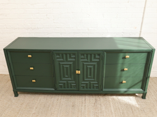 Load image into Gallery viewer, A dark green sideboard cabinet with geometric patterned doors and brass hardware, set against a white brick wall and beige carpet.
