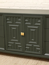Load image into Gallery viewer, A close-up of a dark green cabinet with geometrically patterned doors and a small gold square handle, set against a white brick wall and beige carpet.
