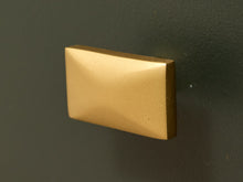 Load image into Gallery viewer, A rectangular gold cabinet knob with a beveled, pyramid-like top edge, mounted on a dark textured wall.
