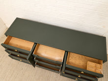 Load image into Gallery viewer, A dark green wooden storage cabinet with multiple drawers pulled open, showing light natural wood interiors, photographed in a room with a white brick wall and beige carpet.
