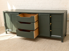 Load image into Gallery viewer, A dark green sideboard cabinet with three open drawers showing light wood interiors, set against a white brick wall and beige carpet.
