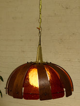 Load image into Gallery viewer, A hanging pendant light with a brass-toned metal frame and multiple curved amber-red glass shades, illuminated against a white brick wall background.
